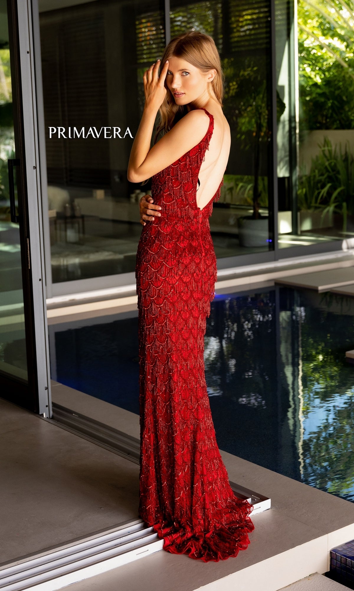 A woman in the Beaded-Fringe Long Primavera Prom Dress 4129—sleeveless, floor-length, and red with beaded details—stands by a glass door near a pool with plants in the background. The word